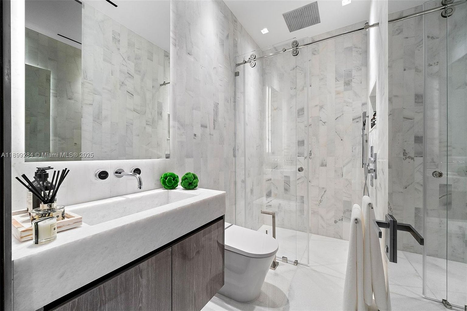 This is a modern primary bathroom featuring marble walls and flooring, a floating vanity with a marble countertop, and a glass-enclosed shower. The bathroom also includes a sleek toilet and contemporary fixtures. The overall impression is luxurious and spa-like.