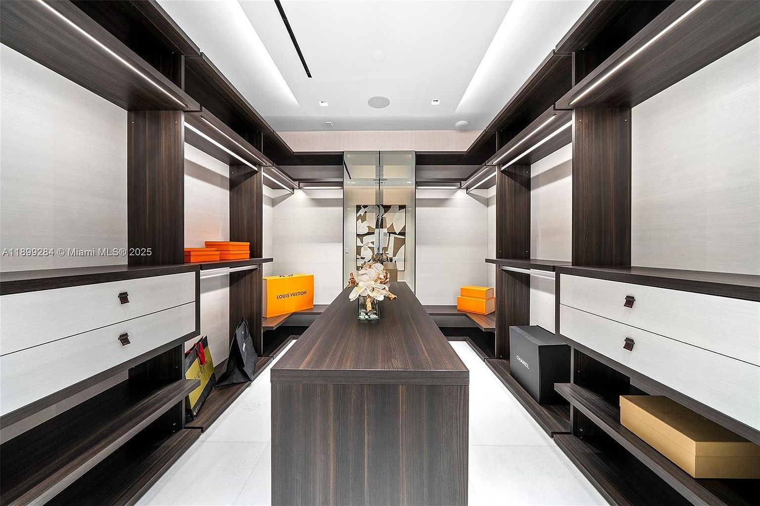 This is a well-organized walk-in closet featuring dark wood shelving and drawers with light-colored fronts. The closet includes ample storage space, with shelves for boxes and hanging rods for clothing. A central island provides additional surface area, and the overall design is modern and luxurious.