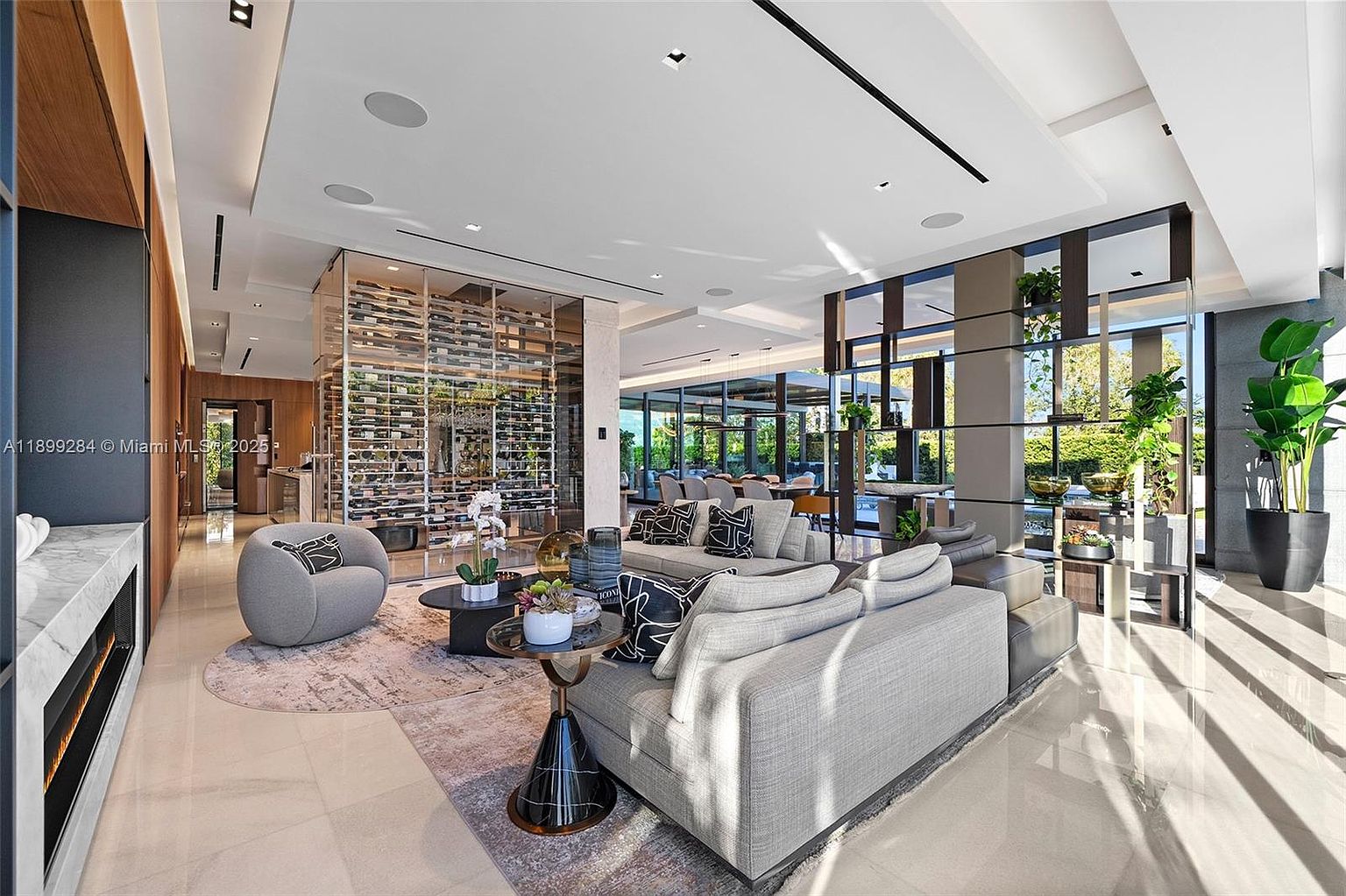 This is a spacious and modern living room featuring a large gray sectional sofa, a glass-enclosed wine cellar, and a decorative shelving unit with plants. The room is well-lit with natural light streaming through large windows, highlighting the polished tile flooring and contemporary design. The perspective is from a wide angle, showcasing the breadth of the living space.
