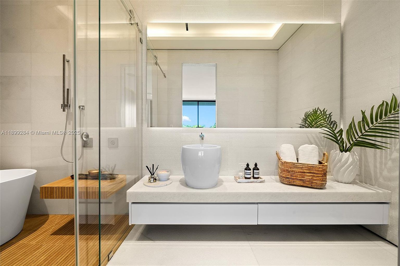 This is a modern primary bathroom featuring a floating vanity with a vessel sink, a large mirror, and a glass-enclosed shower. The color palette is primarily white and neutral, creating a clean and serene atmosphere. The bathroom also includes a wooden bench and teak flooring in the shower area, adding a touch of warmth and luxury.