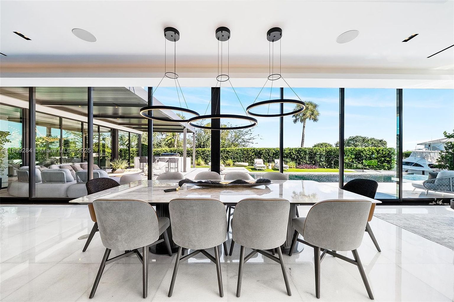 This is a modern dining room featuring a large marble-top dining table surrounded by gray upholstered chairs with dark legs. Three circular pendant lights hang above the table, and floor-to-ceiling windows offer a view of the outdoor pool and landscaping. The room has a clean, contemporary aesthetic with a focus on natural light and open space.