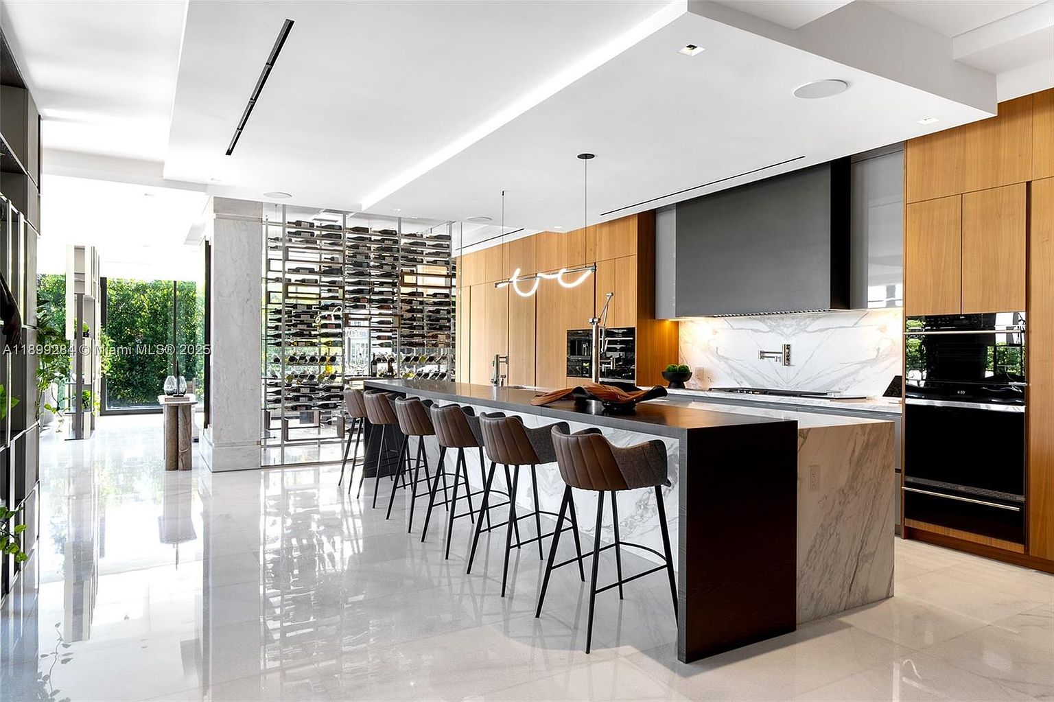 This is a modern, high-end kitchen featuring a large island with seating, sleek wooden cabinetry, and a striking wine rack. The kitchen is well-lit with contemporary lighting fixtures and natural light, highlighting the glossy floors and clean lines. The overall impression is one of luxury and sophistication, perfect for entertaining and gourmet cooking.