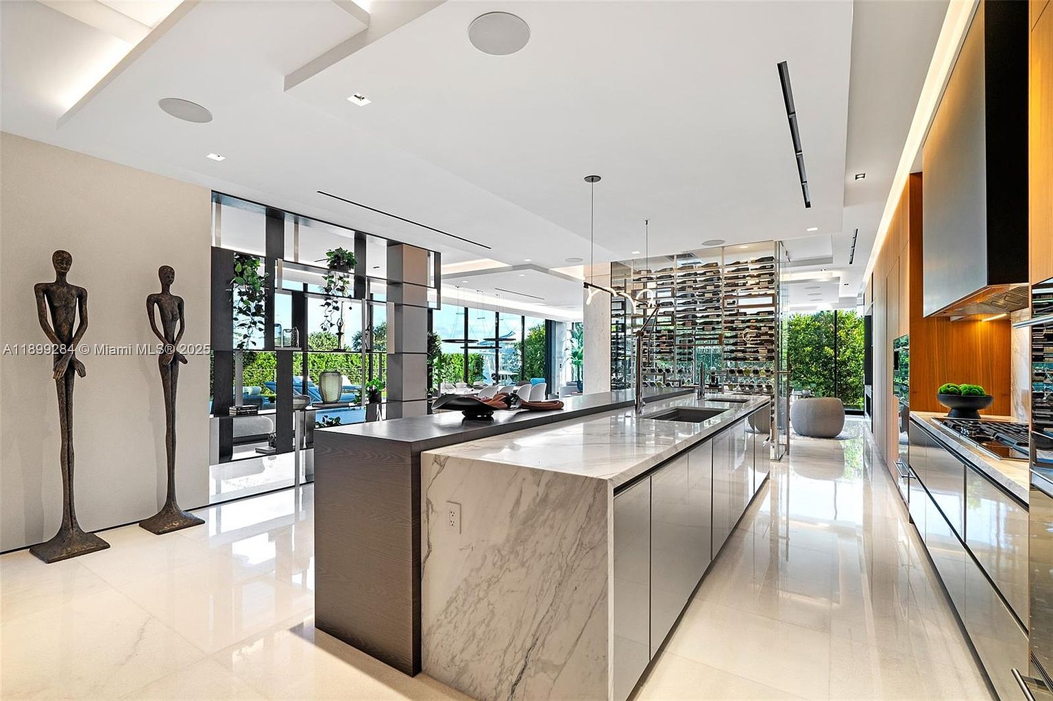 This is a wide-angle shot of a modern, luxurious kitchen featuring a large island with a marble countertop and dark wood accents. The kitchen boasts high-end appliances, a wine rack, and floor-to-ceiling windows that offer a view of the outdoors. Two statues stand near the entrance to the kitchen, adding an artistic touch.