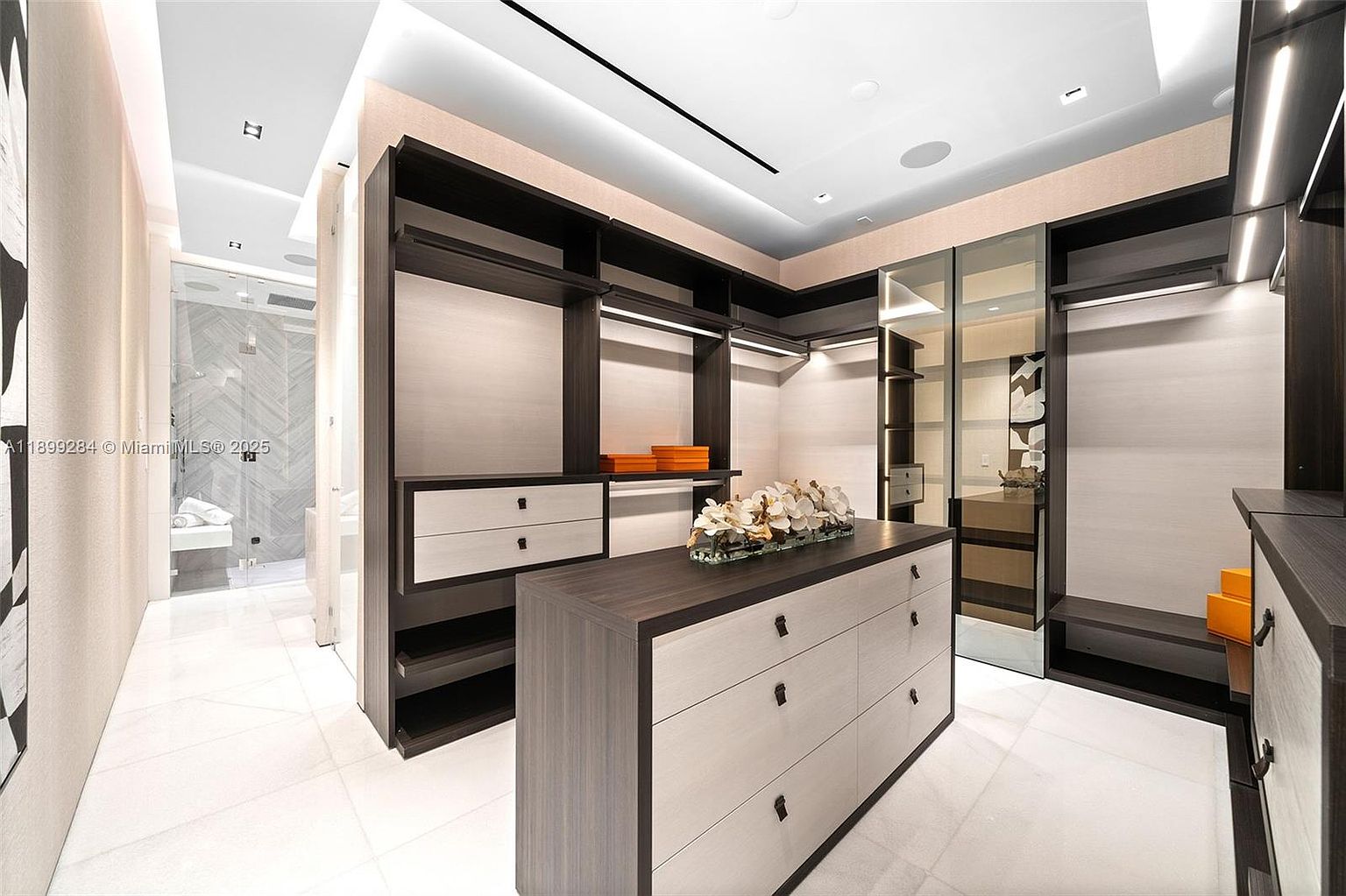 This is a well-organized walk-in closet featuring custom shelving, drawers, and hanging rods. The closet has a neutral color palette with dark wood accents and light-colored walls and drawers. An island in the center provides additional storage and display space, enhancing the luxurious feel of the space.