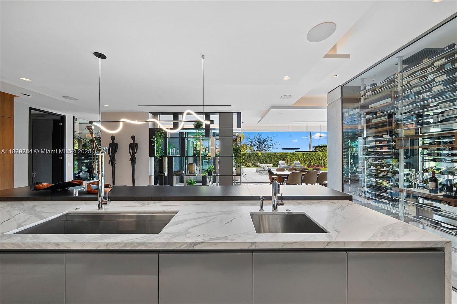 This is a modern kitchen featuring a large island with a marble countertop and dual stainless steel sinks. The kitchen includes sleek gray cabinetry and a glass-enclosed wine cellar. The open layout provides a view of the outdoor dining area and garden, creating a seamless indoor-outdoor living experience.