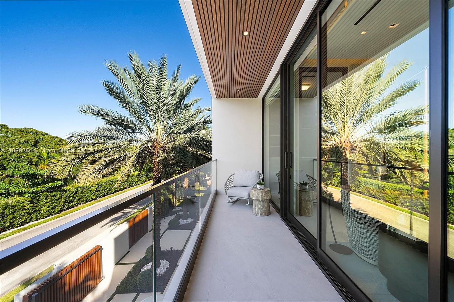 This image showcases a modern balcony with a sleek design. It features glass railings, a comfortable chair, and a small side table. The balcony offers a view of lush greenery and a palm tree, creating a serene and inviting outdoor space. The ceiling is lined with wood adding a touch of warmth.