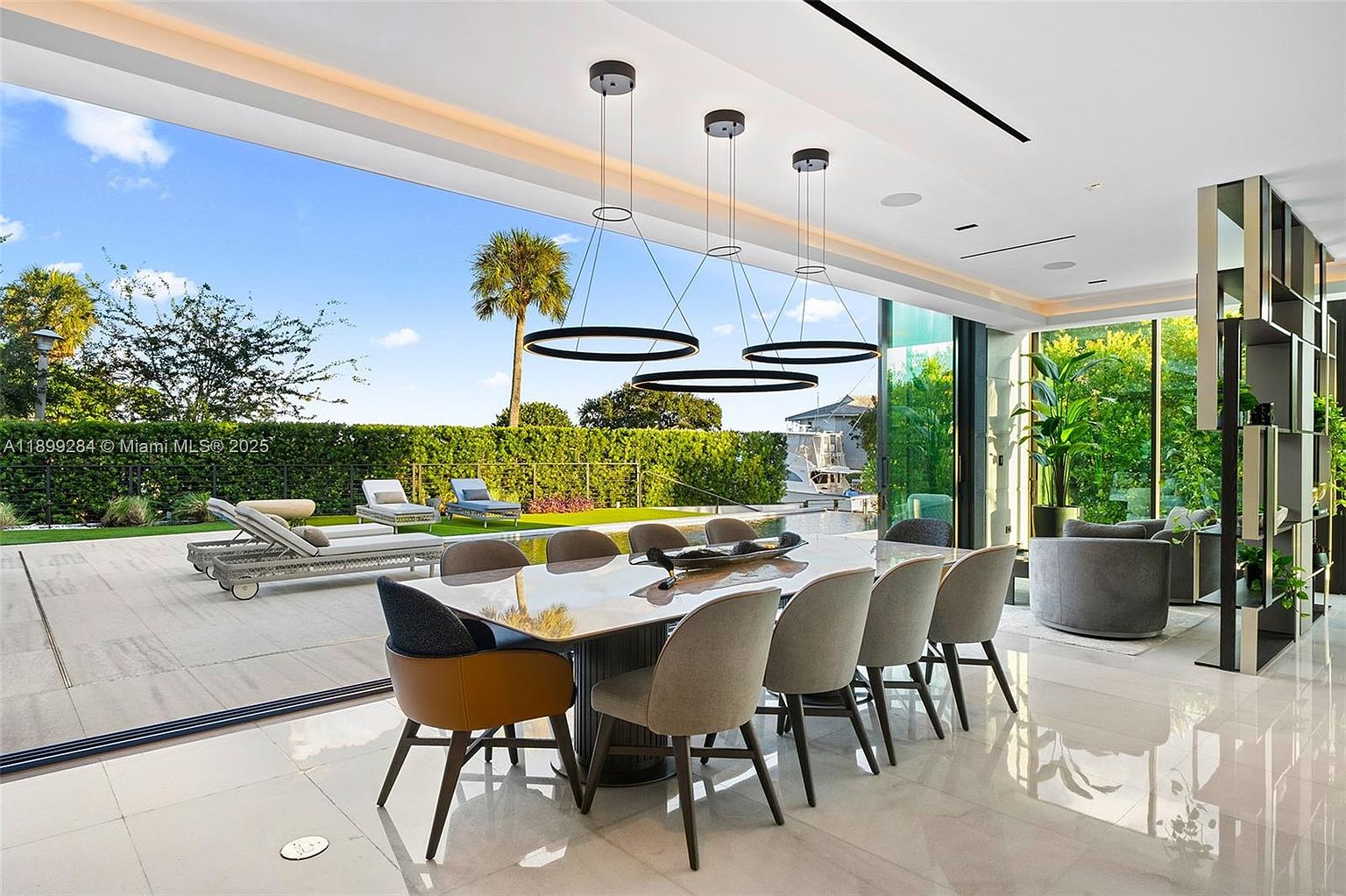 This is a modern dining room featuring a large dining table with seating for eight, illuminated by three circular pendant lights. The room opens to an outdoor patio with lounge chairs and a pool, offering seamless indoor-outdoor living. The interior is bright and airy, with sleek tile flooring and a contemporary design.