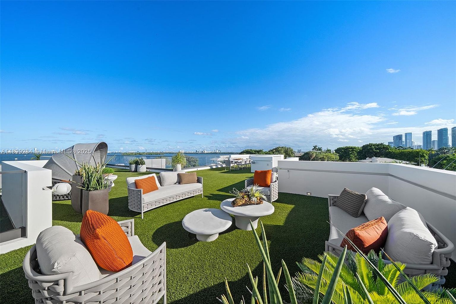 This image showcases a luxurious rooftop patio with modern outdoor furniture, including sofas, chairs, and coffee tables, arranged on artificial turf. The space offers stunning panoramic views of the city skyline and waterfront. The overall impression is one of sophisticated outdoor living and relaxation.