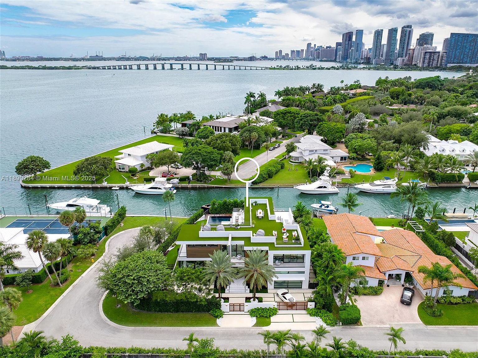 This aerial shot showcases a luxurious waterfront property with a modern architectural design, featuring a flat green roof, a pool, and multiple outdoor seating areas. The property is situated on a canal with boats docked nearby, and the cityscape provides a stunning backdrop, emphasizing the prime location and upscale lifestyle.