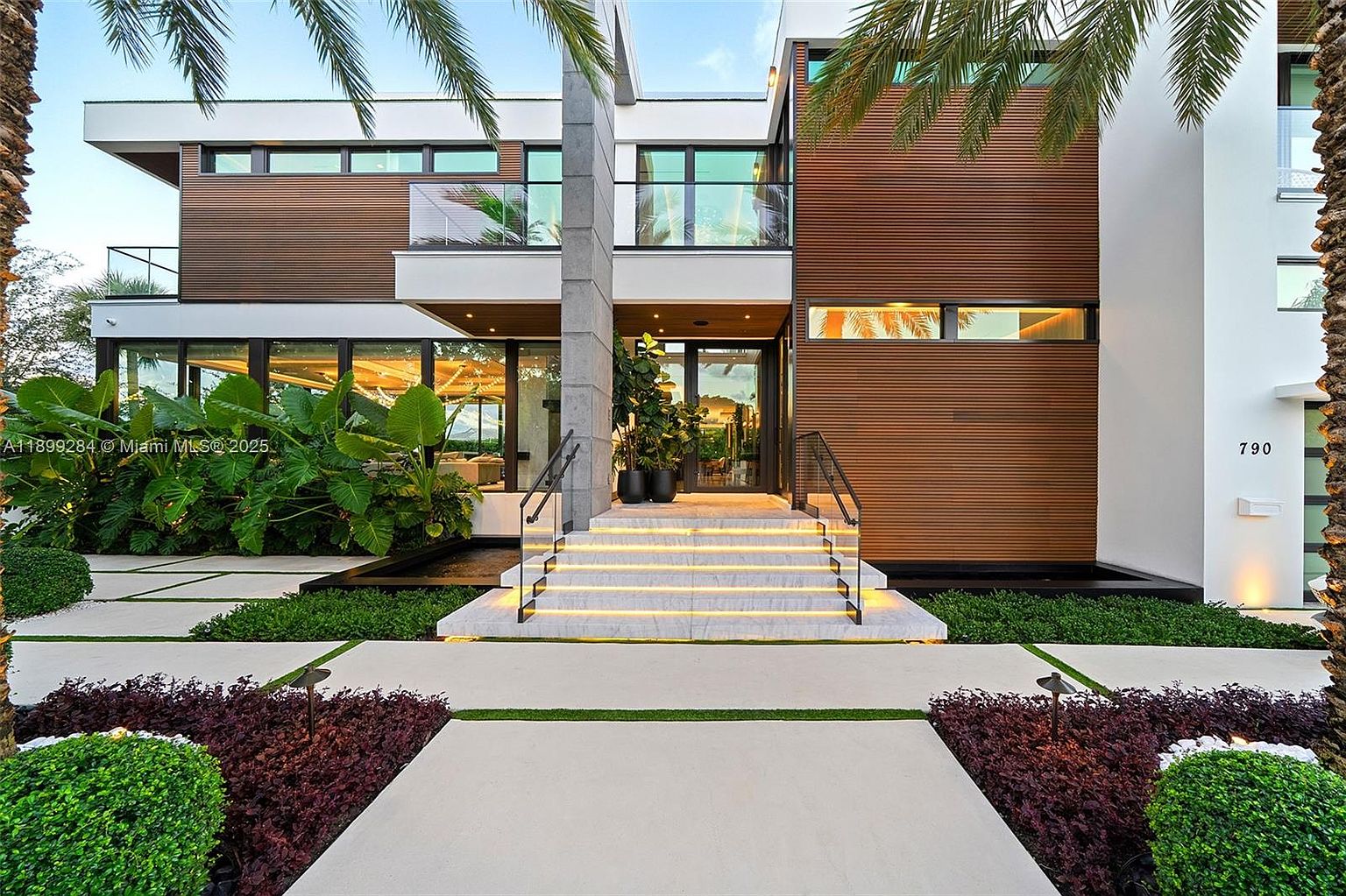 This image showcases the grand entryway of a modern home, featuring a striking facade with a combination of wood paneling, white walls, and a prominent concrete pillar. Illuminated stairs lead to the front door, flanked by lush landscaping and manicured greenery. The overall impression is one of sophisticated design and curb appeal.