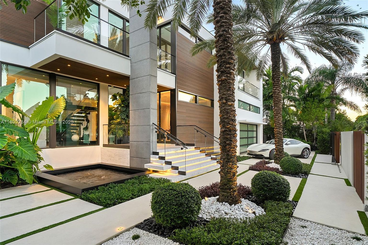 This is a front exterior view of a modern luxury home. The architecture features a combination of white walls, wood paneling, and large glass windows, complemented by a striking water feature and meticulously landscaped garden with palm trees. A white sports car is parked in the driveway, adding to the property's upscale appeal.