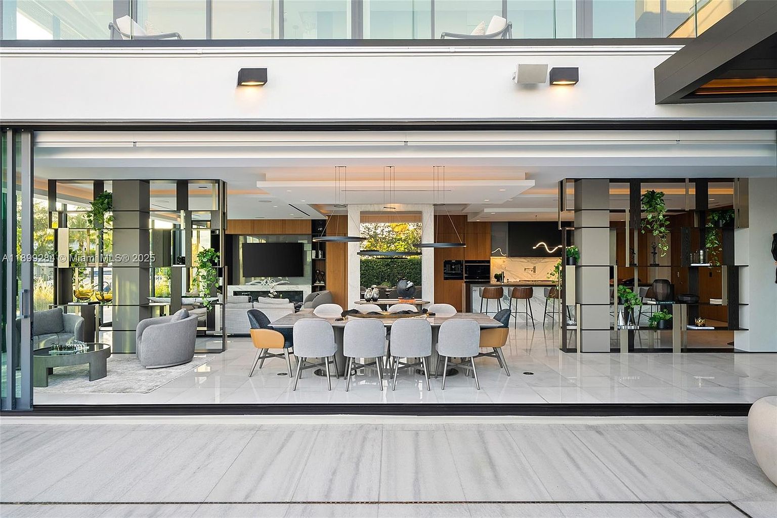 This interior shot showcases a modern dining room seamlessly connected to the living room and kitchen. The dining area features a long wooden table surrounded by contemporary chairs, illuminated by sleek pendant lighting. Large sliding glass doors open to the outside, creating an open and airy atmosphere, while decorative shelving units with plants add a touch of nature to the space.