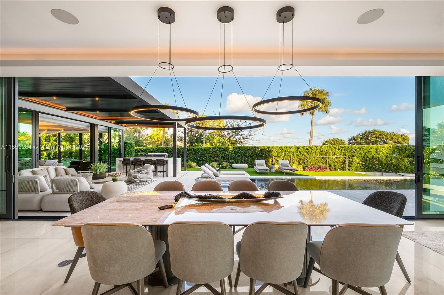This image showcases a luxurious dining room that seamlessly blends indoor and outdoor living. The dining area features a large, elegant table with modern chairs, illuminated by contemporary ring-shaped pendant lights. Sliding glass doors open to a stunning outdoor space with a pool, lounge chairs, and lush landscaping, creating an inviting and sophisticated atmosphere.