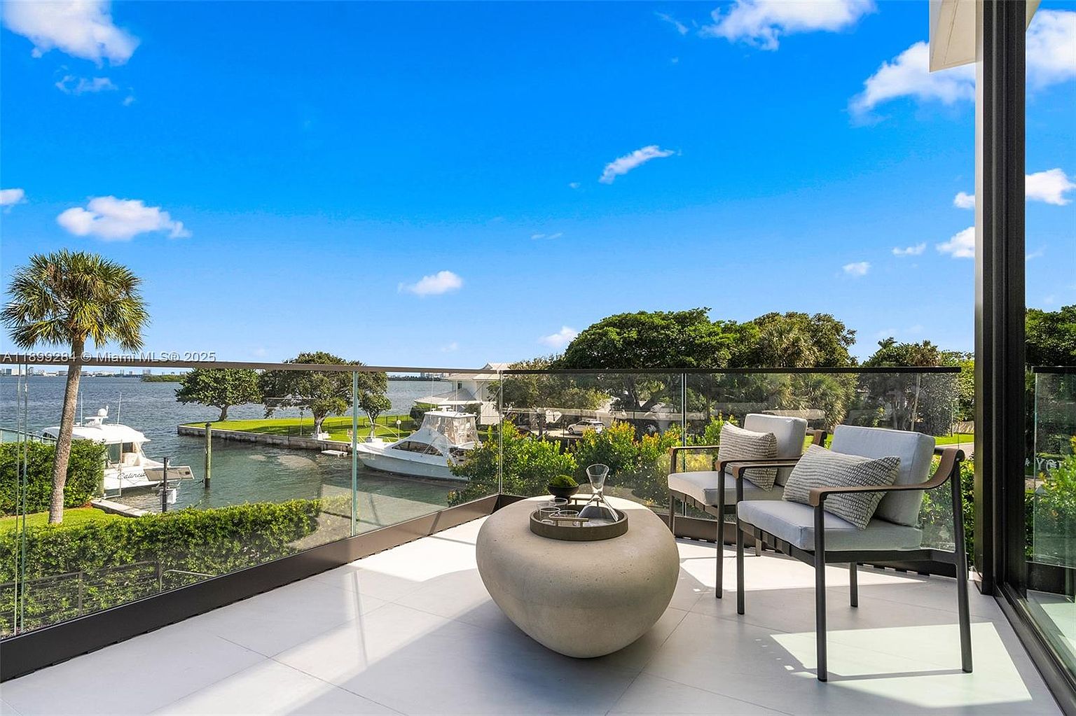 This image showcases a luxurious balcony with a stunning waterfront view. The balcony features modern outdoor furniture, including two chairs and a unique round table, set against a backdrop of lush greenery and boats docked along the canal. The clear glass railing provides an unobstructed view of the water and sky, enhancing the sense of openness and tranquility.