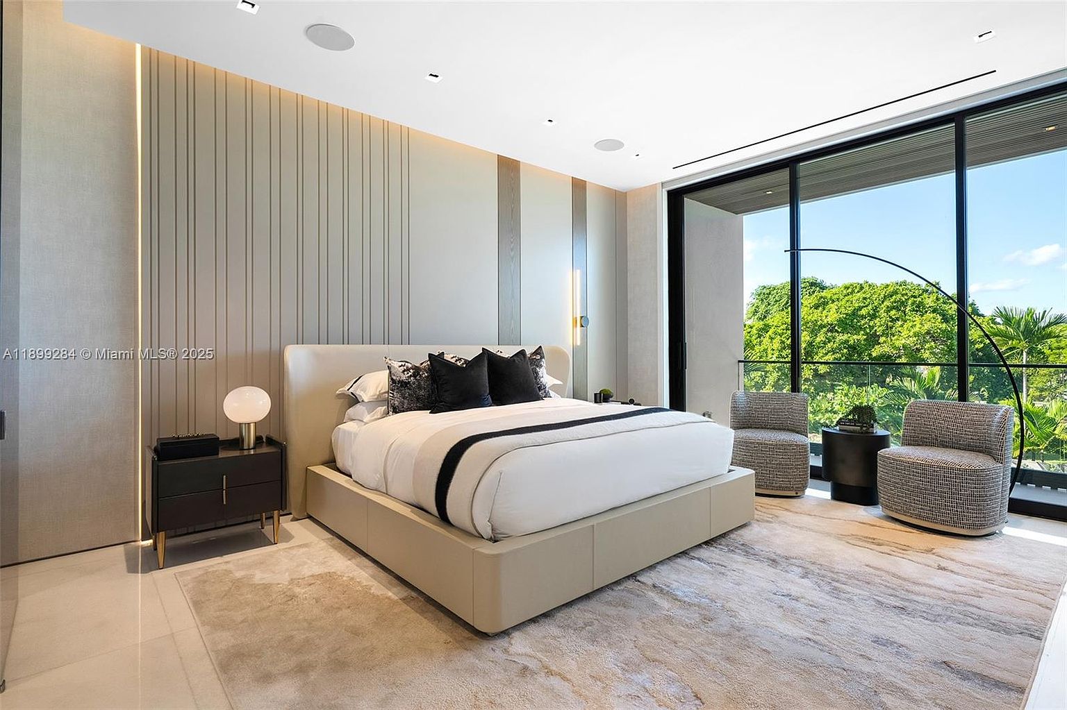 This is a luxurious primary bedroom featuring a large platform bed with a padded headboard, flanked by a modern nightstand with a table lamp. The room boasts a neutral color palette with textured wall panels and a large window offering a view of lush greenery. Two armchairs are placed near the window, creating a cozy seating area.