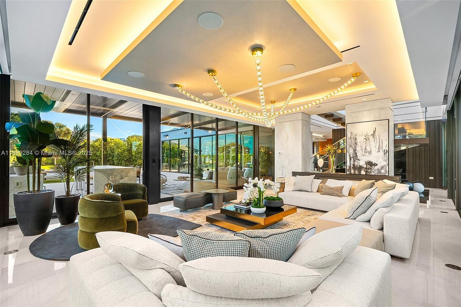 This is a luxurious living room featuring a modern design with large windows offering an outdoor view. The room is furnished with multiple sofas, a unique chandelier, and stylish decor. The open floor plan and high ceilings create a spacious and elegant atmosphere, perfect for entertaining or relaxing.