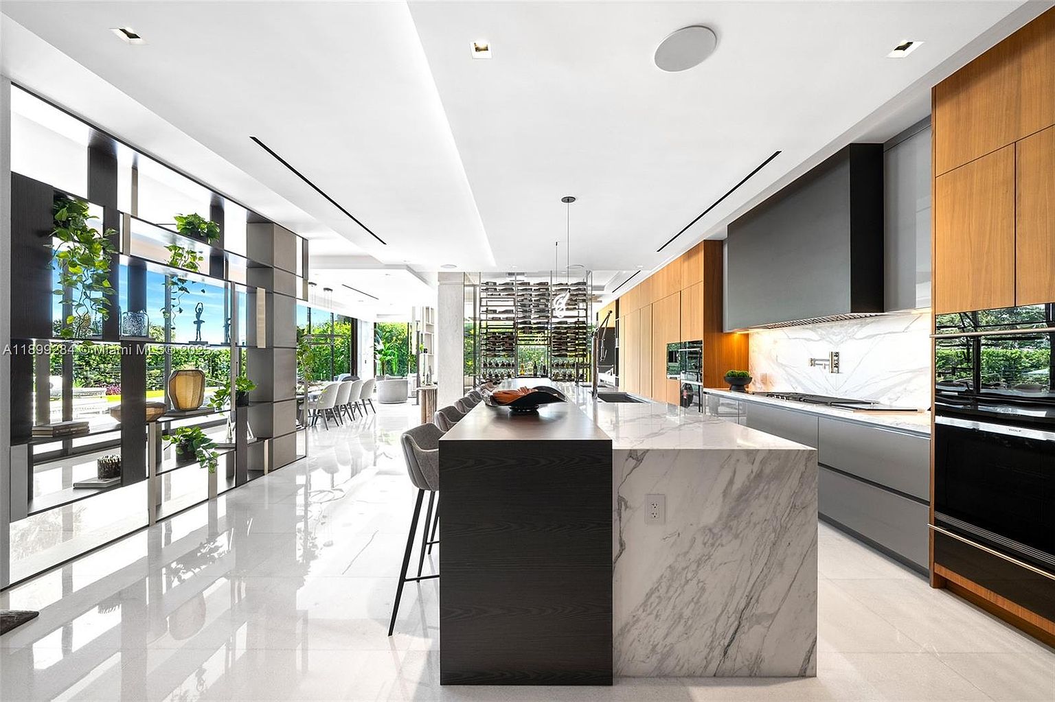 This is a modern, high-end kitchen featuring a large island with a two-tone design, combining dark wood and marble. The kitchen is equipped with sleek, built-in appliances and custom cabinetry, complemented by a marble backsplash. The open layout and reflective flooring create a spacious and luxurious feel, enhanced by natural light streaming in from the adjacent dining area.