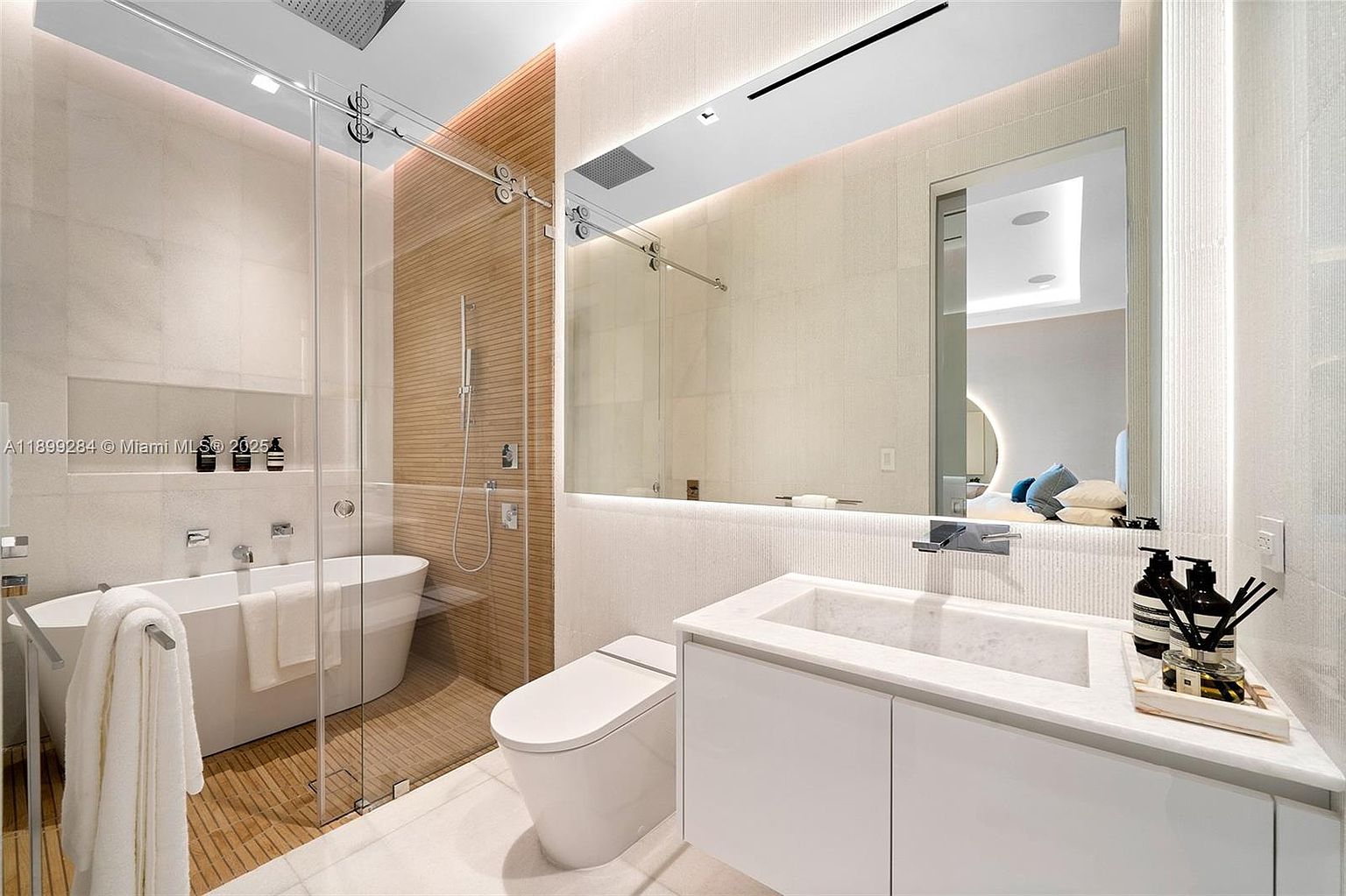 This is a modern primary bathroom featuring a freestanding bathtub enclosed by a glass shower, a wall-mounted toilet, and a sleek vanity with a marble countertop and integrated sink. The bathroom is illuminated by recessed lighting and a large mirror with integrated lighting, creating a bright and luxurious atmosphere. The color palette is neutral, with white and beige tones dominating the space.