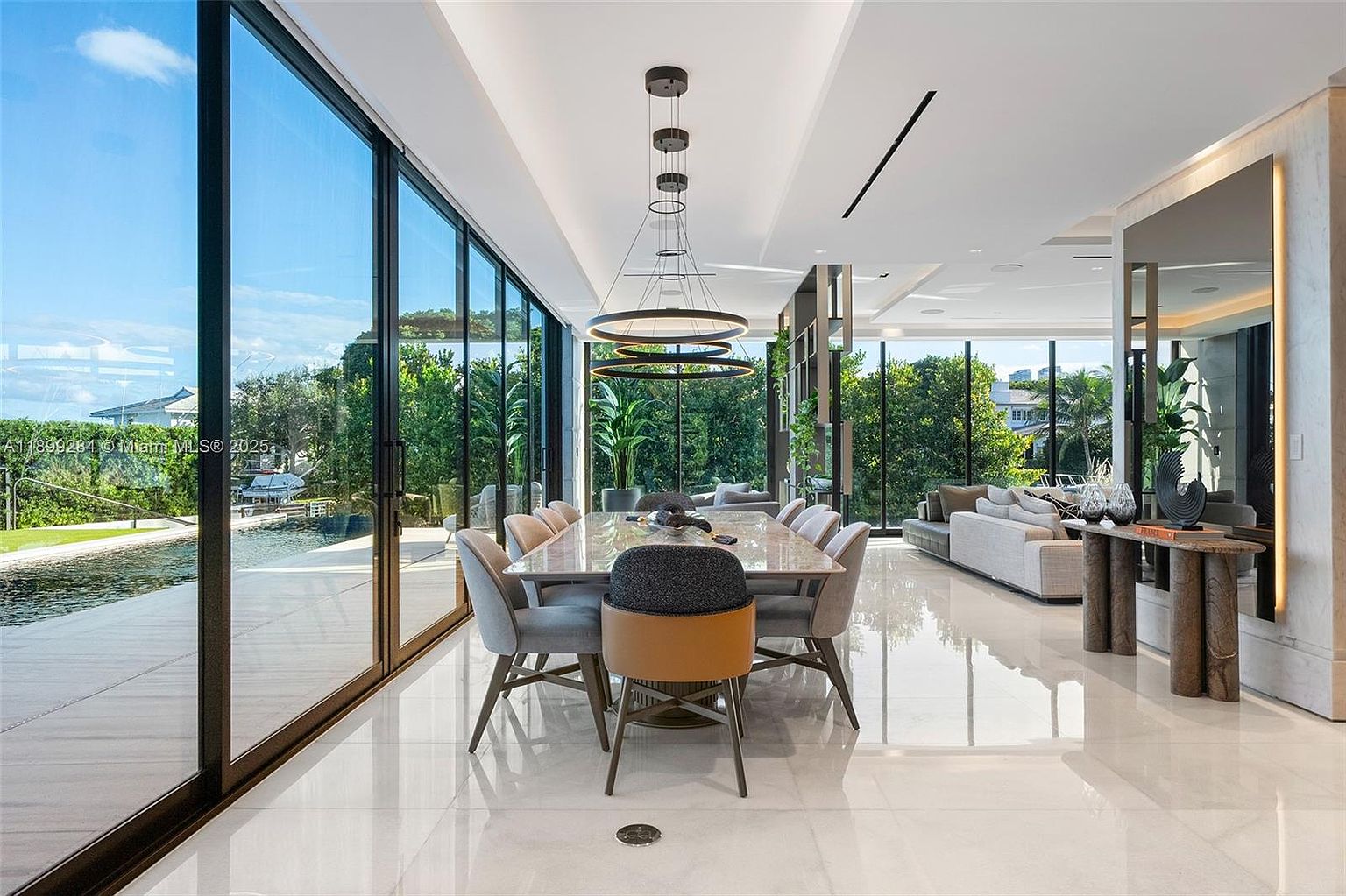 This interior shot showcases a luxurious dining room with a long marble table surrounded by modern chairs. A striking circular chandelier hangs above the table, and floor-to-ceiling windows offer views of the lush exterior. The polished marble floors and minimalist decor contribute to an elegant and spacious atmosphere, perfect for upscale living.