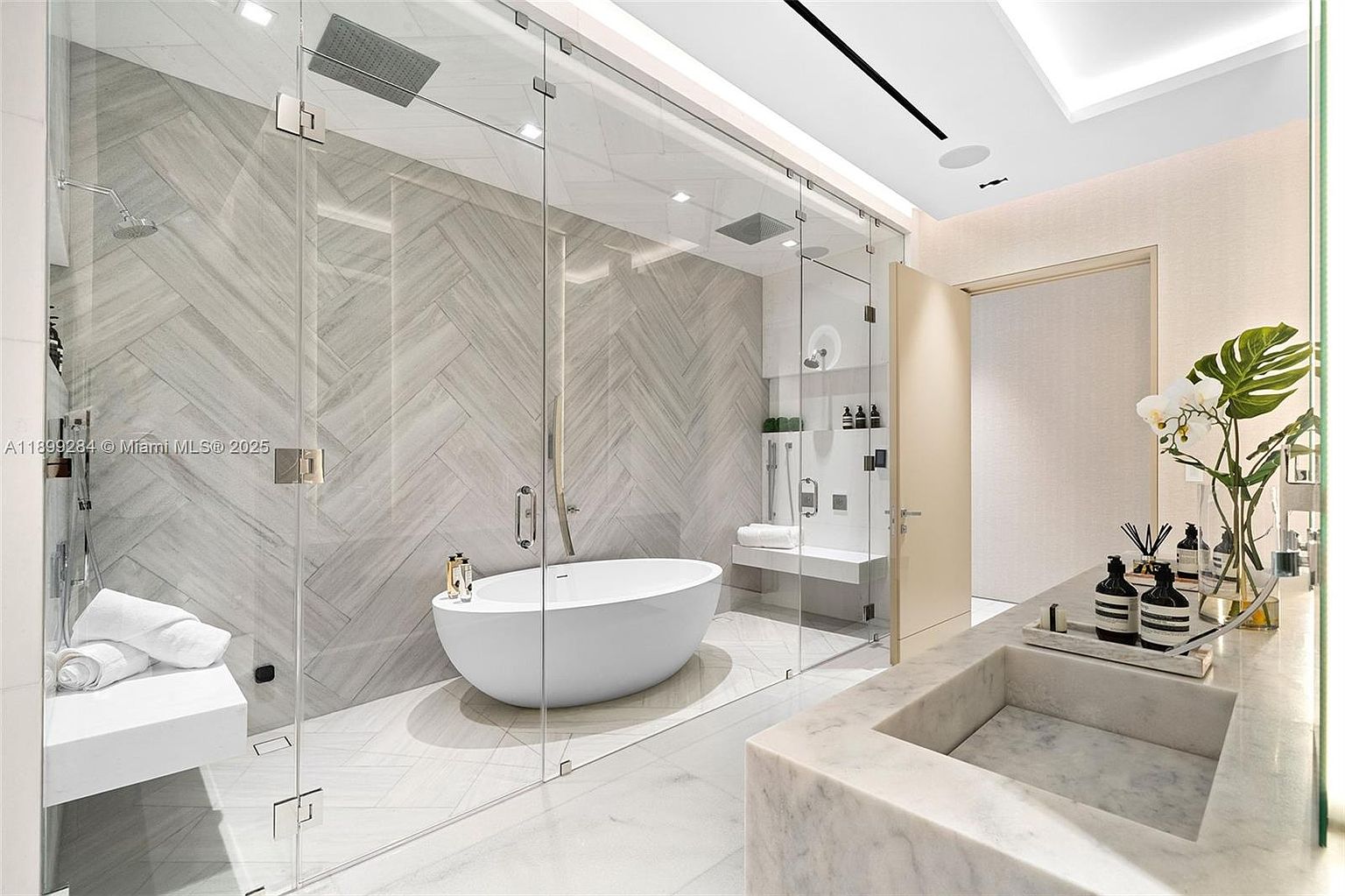 This is a luxurious primary bathroom featuring a modern freestanding bathtub inside a glass-enclosed shower area with dual shower heads. The bathroom boasts marble countertops, a built-in sink, and stylish decor, creating a spa-like atmosphere. The design is clean, contemporary, and focused on relaxation.