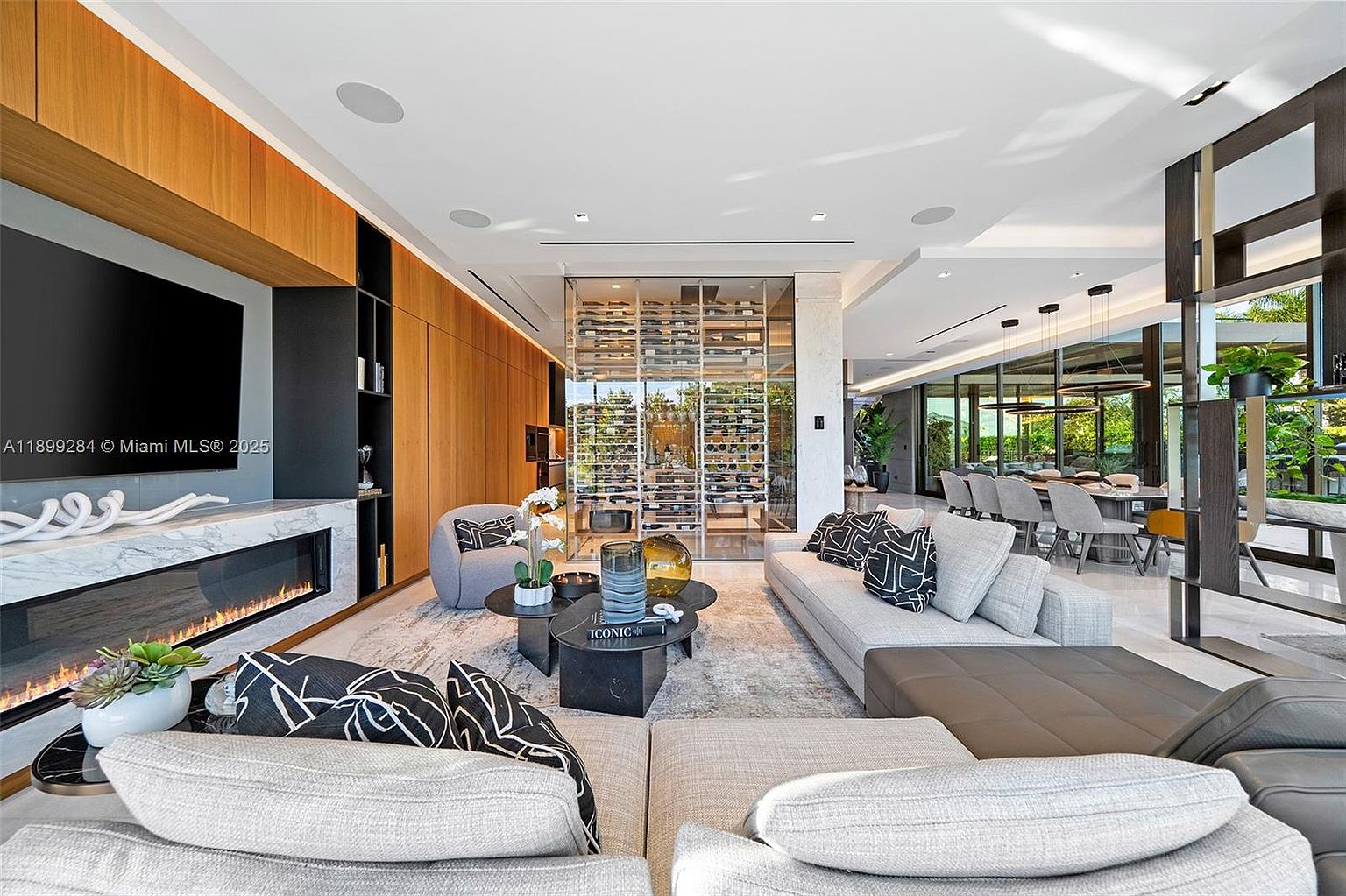 This is a luxurious living room featuring a modern fireplace with a marble surround and a large flat-screen TV. The room is furnished with comfortable sofas and stylish coffee tables, complemented by a glass-enclosed wine cellar in the background. The open-concept design flows into the dining area, creating a spacious and elegant living space.
