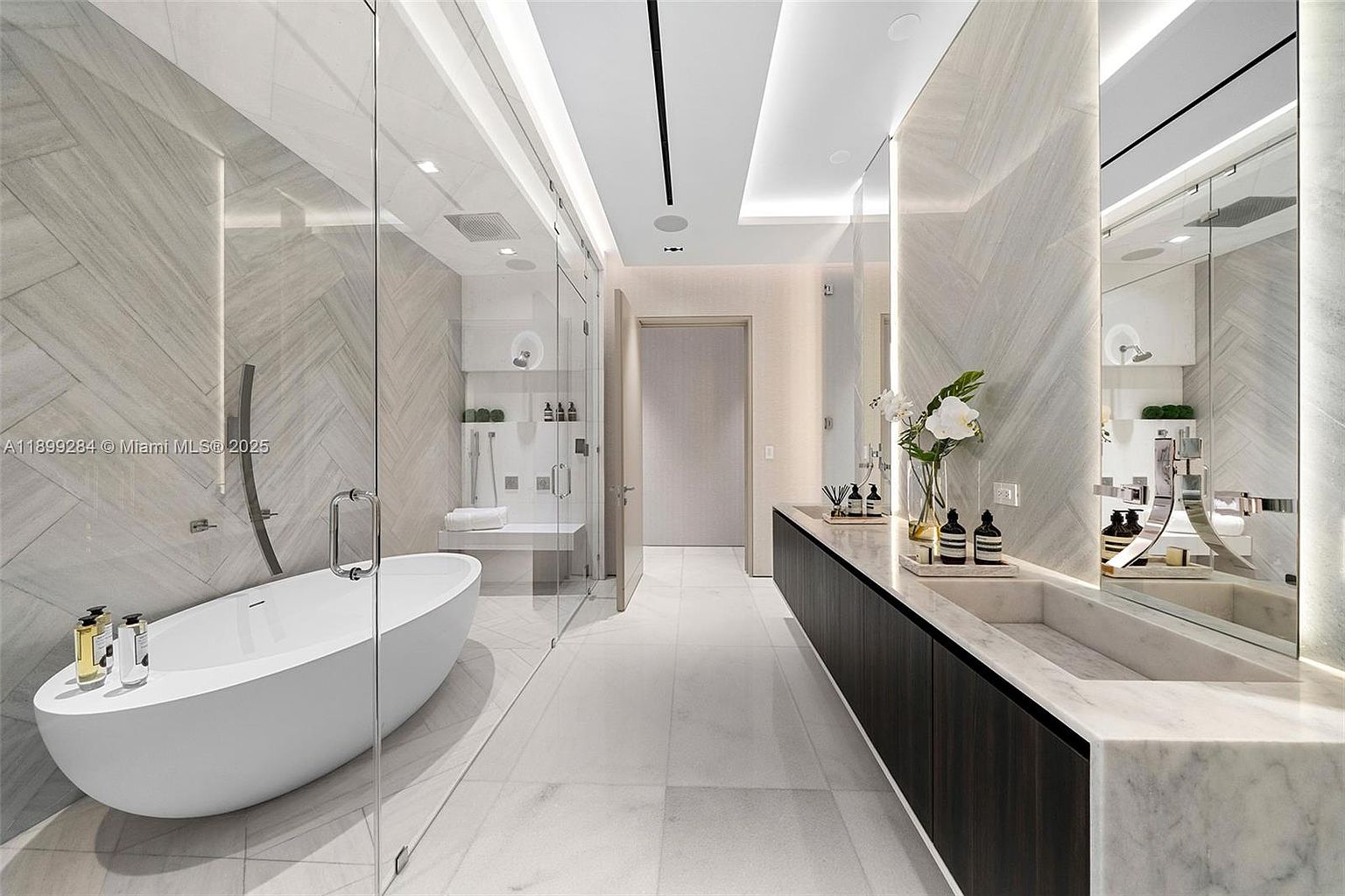 This is a luxurious primary bathroom featuring a freestanding white bathtub, a glass-enclosed shower, and a long vanity with dark wood cabinetry and a marble countertop. The walls are adorned with light-colored herringbone tile, and the space is illuminated by recessed lighting and integrated mirror lighting. The overall impression is modern, clean, and spa-like.