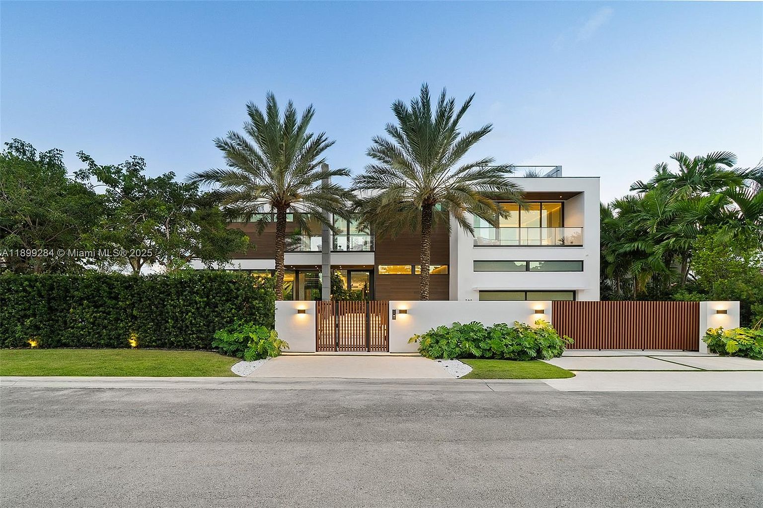 This is a front exterior view of a modern two-story house. The house features a combination of white and wood paneling, with large windows and a balcony. Two tall palm trees stand in front of the house, adding to the tropical feel. A gated entrance and well-maintained landscaping enhance the property's curb appeal.