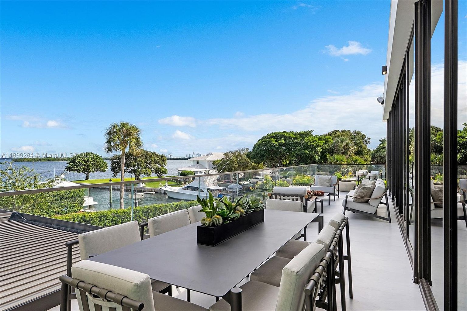 This image showcases a luxurious outdoor patio or balcony area, featuring a large dining table with seating for eight, complemented by comfortable lounge furniture. The space is surrounded by a glass railing, offering unobstructed views of the waterfront, including boats and lush greenery. The overall impression is one of sophisticated outdoor living and relaxation.
