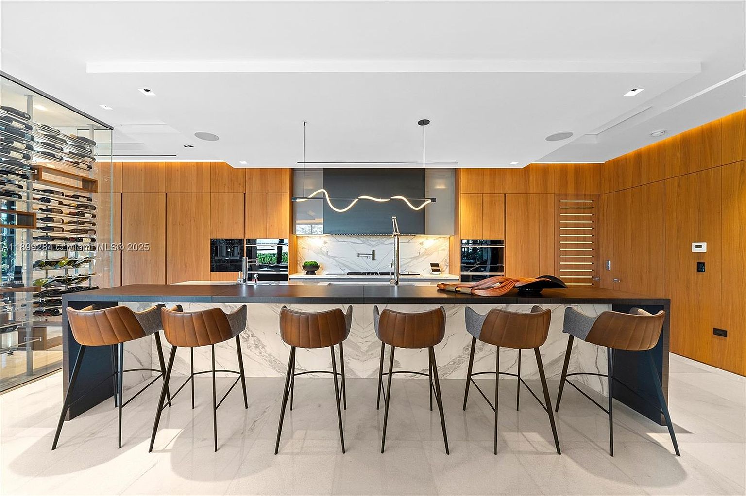 This is a modern kitchen featuring sleek wood cabinetry, a dark countertop island with seating, and marble-like accents. The kitchen is well-lit with modern lighting fixtures and recessed lighting, creating a warm and inviting atmosphere. A wine cellar is visible on the left, adding a touch of luxury.