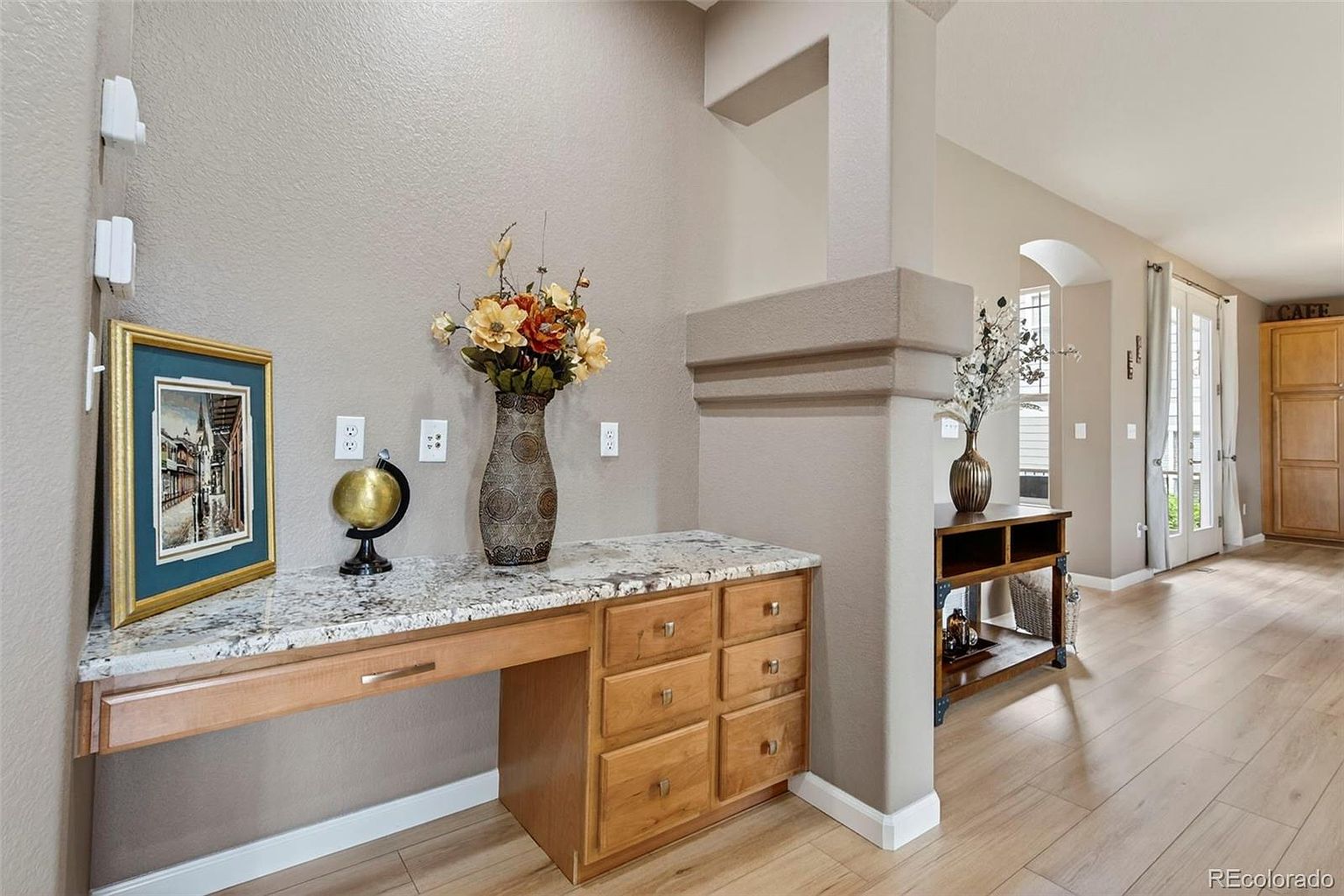 This interior shot showcases a hallway area with a built-in desk featuring granite countertops and wooden drawers. A decorative vase with flowers and a framed picture add a touch of elegance. The flooring is light wood, and the overall impression is clean and well-maintained, suggesting a functional and stylish space.