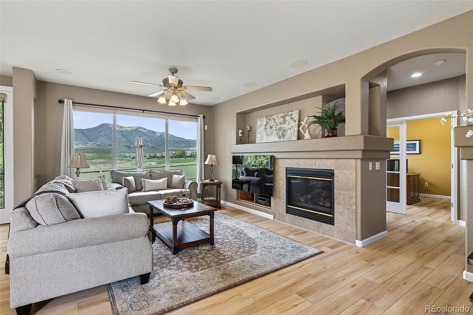 This is a well-lit living room featuring a comfortable gray sofa set, a wooden coffee table on a patterned rug, and a fireplace with a mounted television above it. Large windows offer a scenic view of mountains, and the room has hardwood flooring, creating a warm and inviting atmosphere. The perspective is from the entrance of the room, showcasing the entire space.