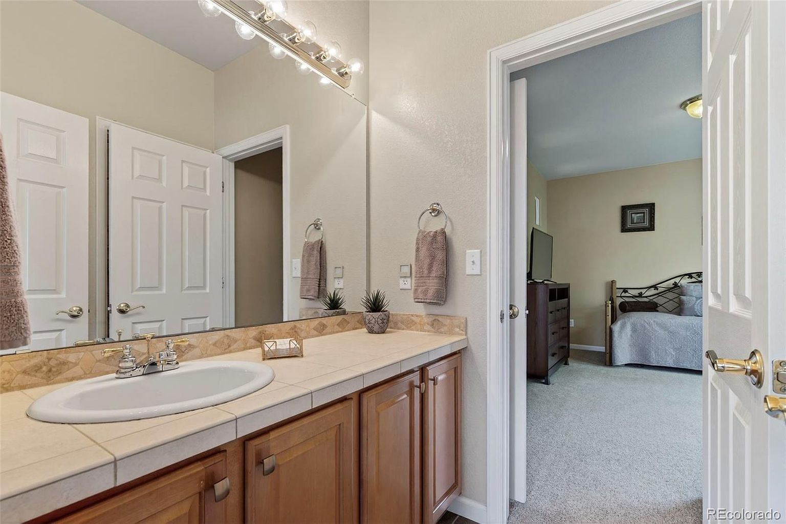 This is a bathroom featuring a vanity with a sink, a tiled countertop, and wooden cabinets. A large mirror hangs above the vanity, reflecting a nearby door and a glimpse into an adjacent bedroom. The bathroom appears clean and well-maintained, with neutral wall colors and simple decor.