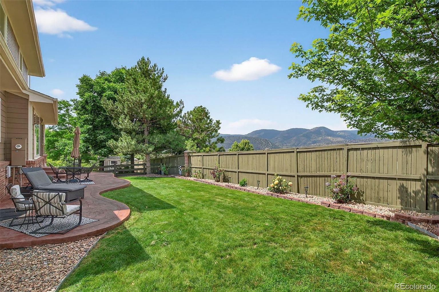 This image showcases a well-maintained backyard with a lush green lawn, a wooden fence, and a paved patio area with outdoor furniture. The yard features landscaping with flowers and trees, and mountains are visible in the background. The overall impression is a serene and inviting outdoor space, perfect for relaxation and entertainment.