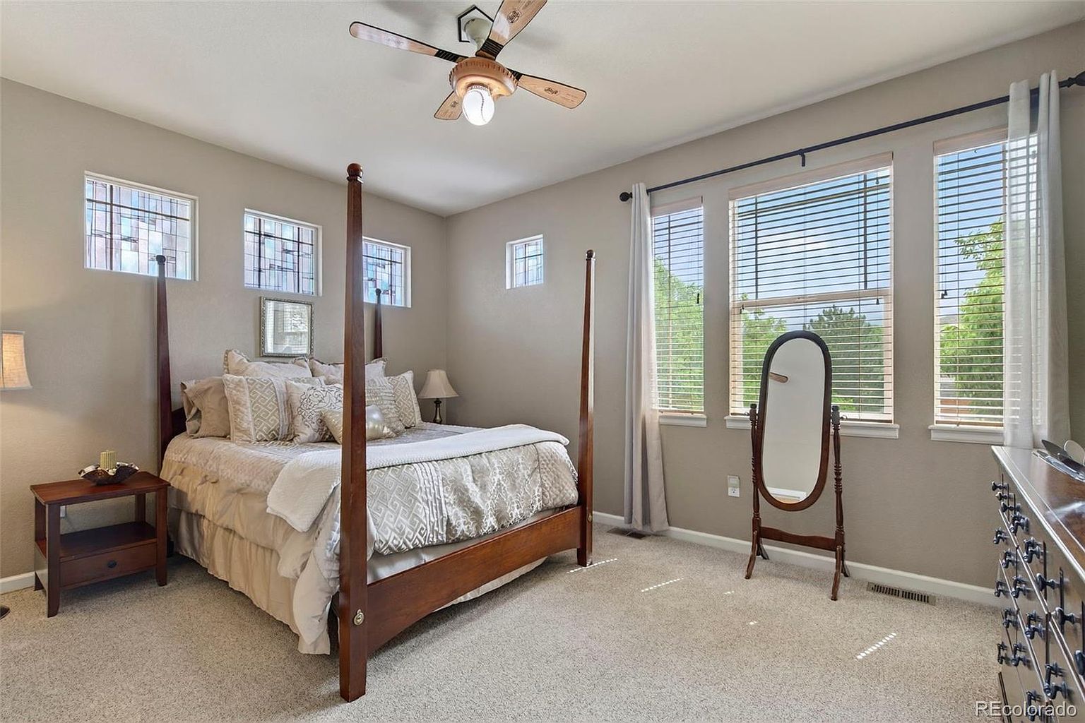 This is a primary bedroom featuring a four-poster bed with decorative pillows and a patterned comforter. The room has neutral-colored walls, carpeted flooring, and multiple windows with blinds and curtains, providing natural light. A wooden side table and a full-length mirror add to the room's functionality and style.