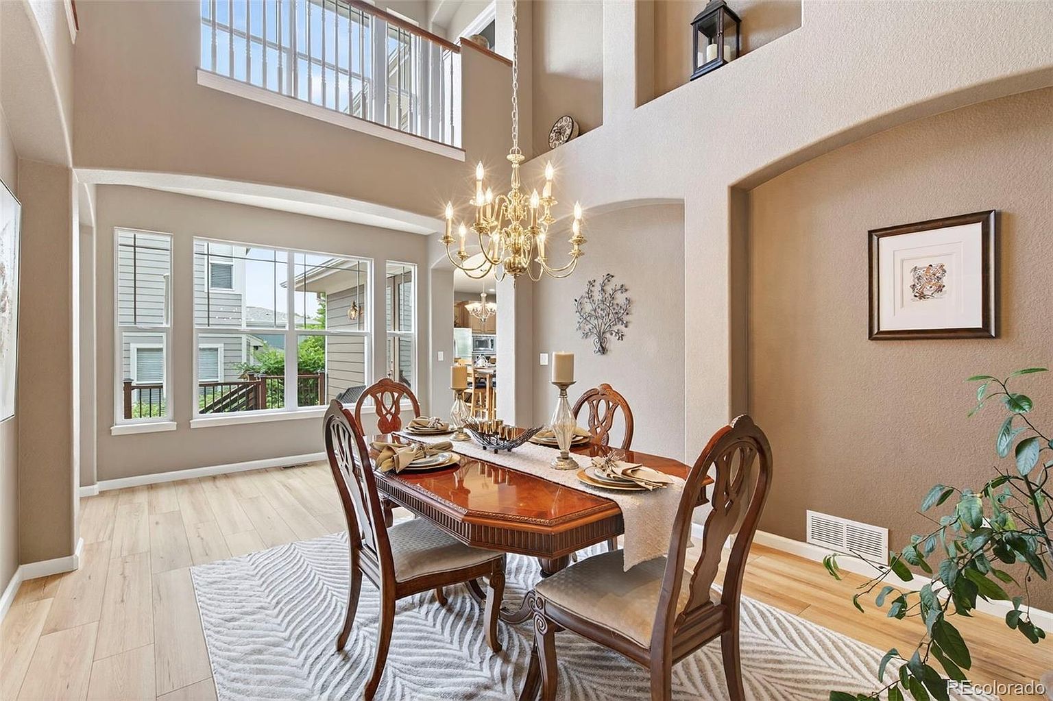 This is an interior shot of a dining room featuring a wooden dining table set for a meal, surrounded by four chairs on a patterned rug. A chandelier hangs above the table, and natural light floods the room through a large window. The walls are painted in neutral tones, and there is a framed picture on the wall, creating a warm and inviting atmosphere.