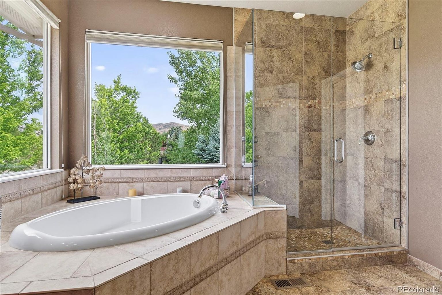 This is a luxurious primary bathroom featuring a large soaking tub situated beneath a wide window offering a view of lush greenery. A glass-enclosed shower with tiled walls and a mosaic accent strip adds to the spa-like atmosphere. The bathroom is finished with neutral-toned tiles and fixtures, creating a serene and elegant space.