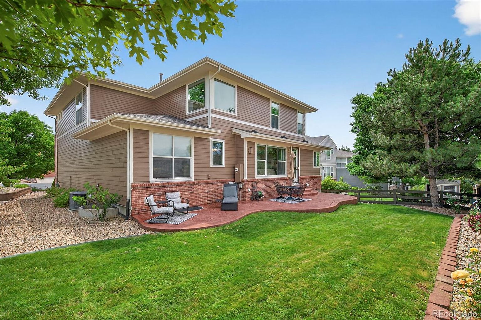 The image showcases the rear exterior of a two-story house with brown siding and a brick foundation. A curved red brick patio features outdoor seating, and a well-maintained green lawn extends to a brick-lined flower bed. Mature trees and a wooden fence add to the property's appeal, creating a serene backyard setting.