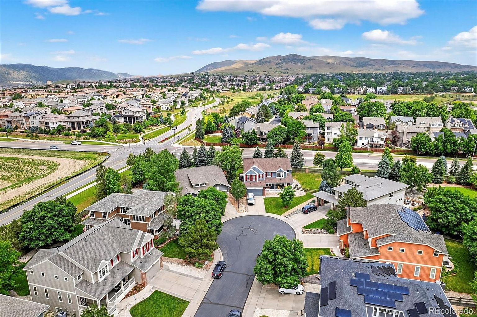 This aerial view showcases a well-planned residential neighborhood with neatly arranged houses, manicured lawns, and mature trees. The homes feature a variety of architectural styles, some with solar panels, and are connected by a network of streets and cul-de-sacs. In the background, rolling hills and mountains provide a scenic backdrop, enhancing the desirability of this suburban community.