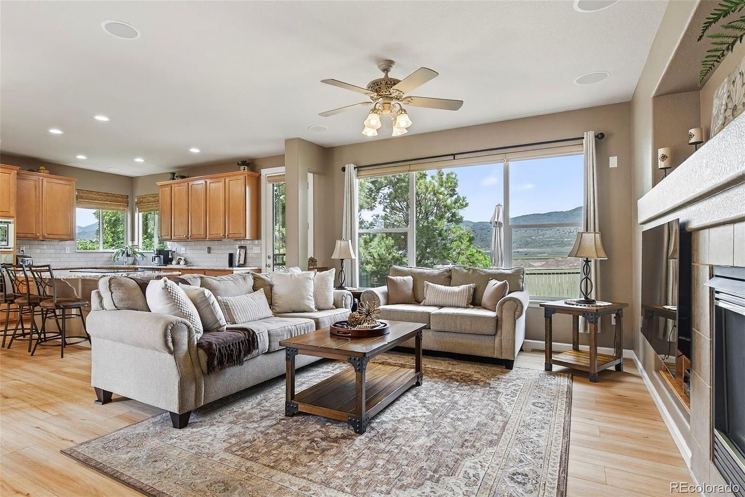 This is an interior shot of a living room featuring two sofas, a wooden coffee table, and a large area rug. The room has a neutral color palette with light wood flooring and a ceiling fan. Large windows provide natural light and a view of the outdoors, while a fireplace adds a cozy touch.