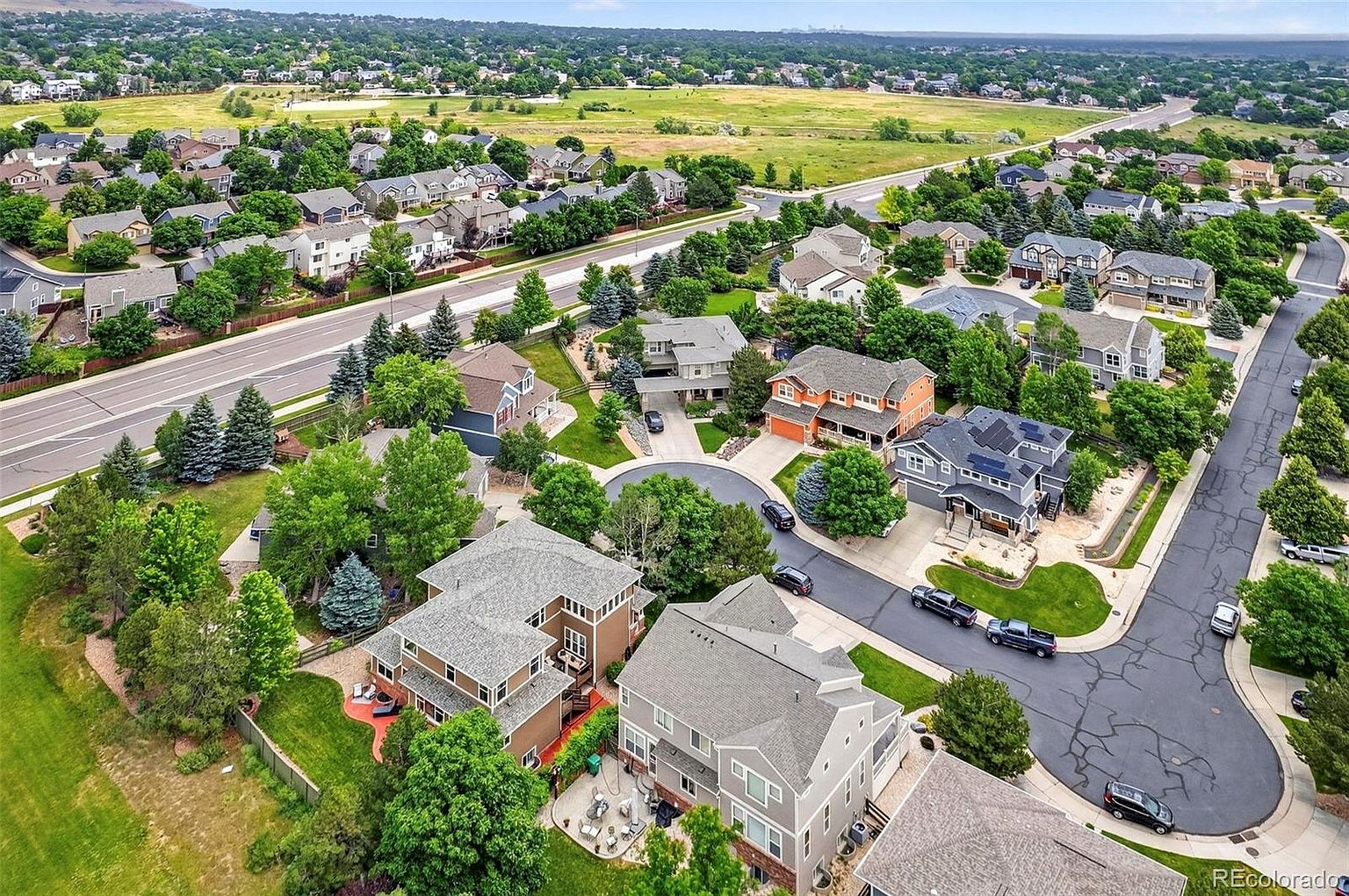 This aerial shot showcases a well-planned residential neighborhood with neatly arranged houses, lush green lawns, and mature trees. The homes feature various architectural styles, some with solar panels, and are connected by paved streets with cul-de-sacs. A large green field borders the neighborhood, providing a sense of open space and tranquility.