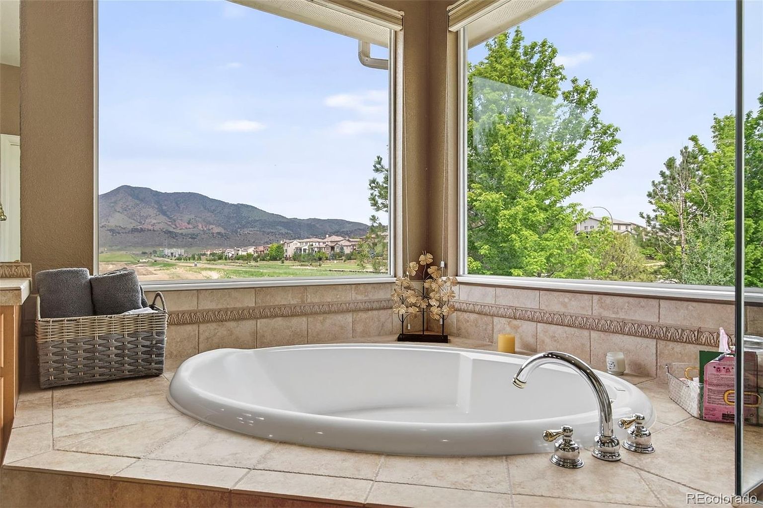 This is a luxurious primary bathroom featuring a large, oval-shaped soaking tub positioned in front of a large window with views of mountains and greenery. The bathroom is tiled in neutral tones, and the tub is complemented by chrome fixtures. A basket of towels sits nearby, adding a touch of comfort and convenience.