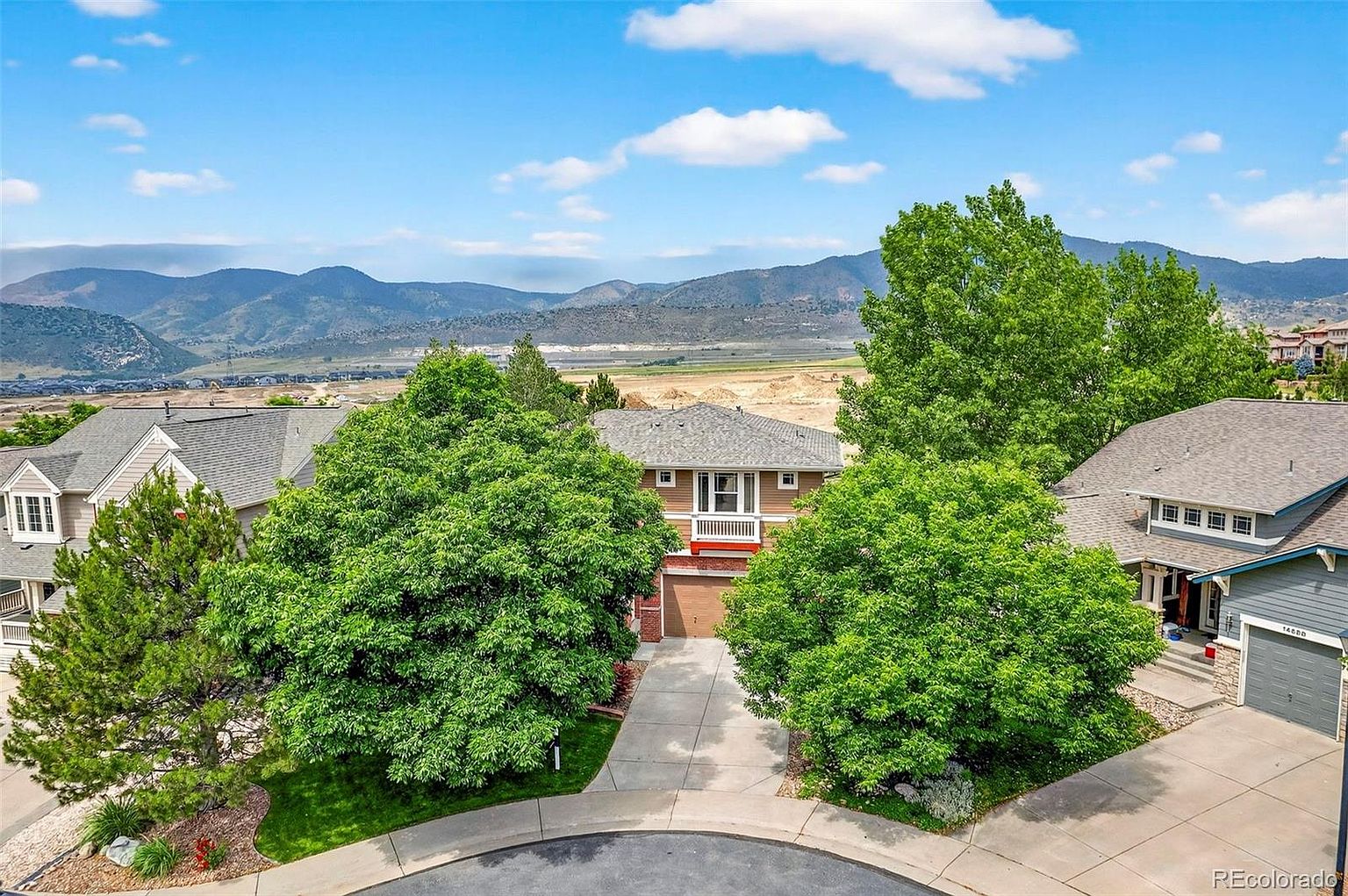 This aerial view showcases a well-maintained suburban home nestled among mature trees. The property features a neatly manicured lawn, a paved driveway leading to a garage, and a charming balcony. In the background, rolling hills and a clear blue sky with scattered clouds create a picturesque setting, enhancing the property's appeal.