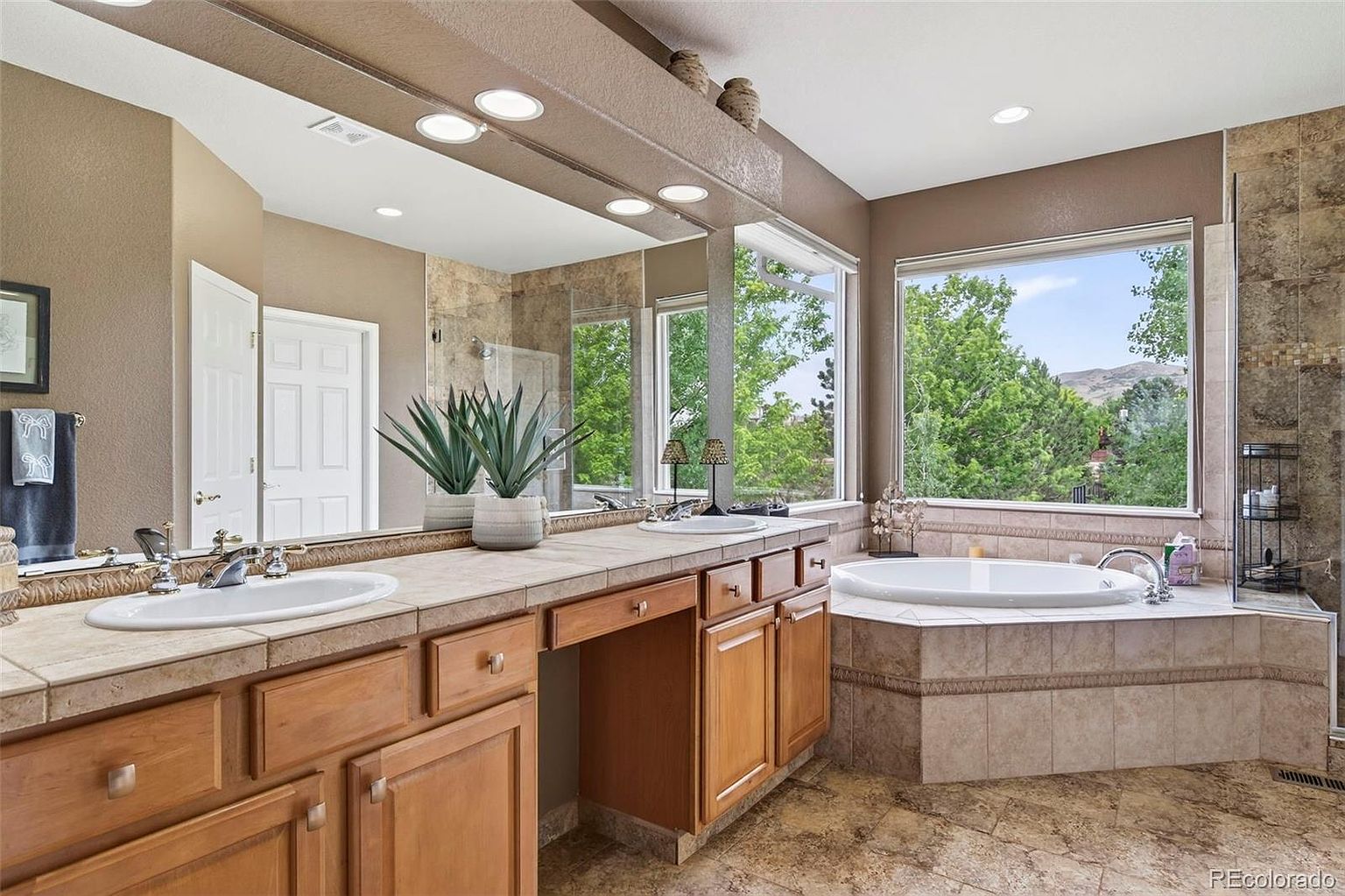 This is a primary bathroom featuring a double vanity with wooden cabinets and tile countertops. A large mirror spans the length of the vanity, reflecting natural light from the adjacent windows overlooking lush greenery. A soaking tub is situated near the windows, and the bathroom is finished with neutral-toned tile flooring and walls, creating a serene and spa-like atmosphere.