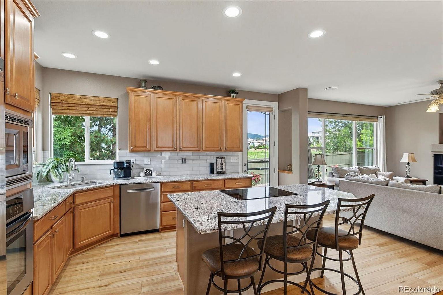 This is a well-lit kitchen featuring wooden cabinetry, granite countertops, and stainless steel appliances. A central island with bar seating provides a focal point, while large windows offer views of the outdoors. The open layout seamlessly connects the kitchen to the living area, creating a spacious and inviting atmosphere.