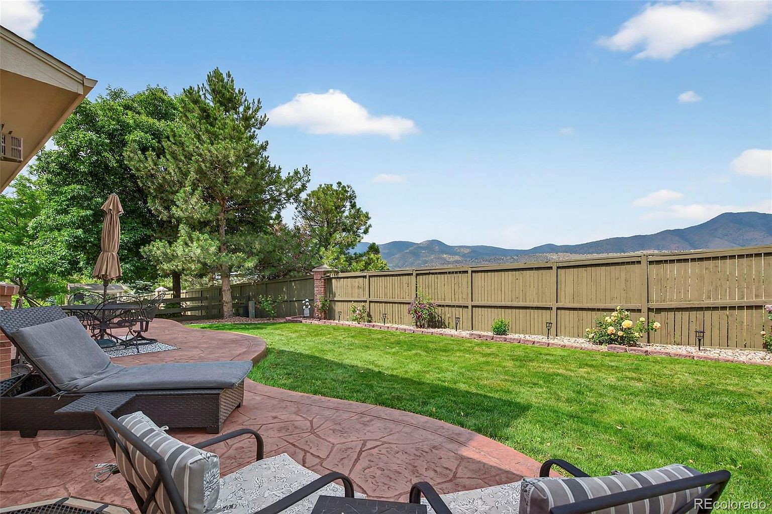 This image showcases a well-maintained backyard featuring a stamped concrete patio with outdoor seating, a lush green lawn, and a wooden fence providing privacy. The yard is landscaped with small plants and flowers along the fence line, and mountains are visible in the distance. The scene conveys a sense of tranquility and outdoor living.