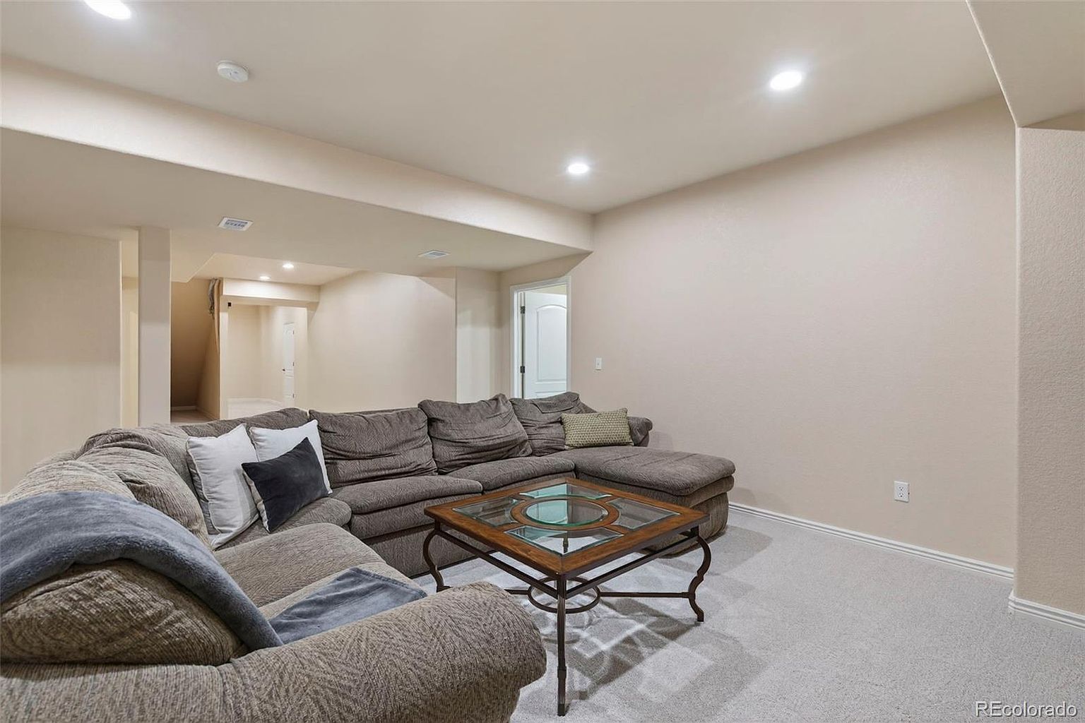 This is an interior shot of a living room featuring a large sectional sofa in neutral tones, accented with throw pillows and a blanket. A glass-topped coffee table sits in the center of the room, and the walls are painted in a light, warm color. The room is well-lit with recessed lighting, creating a cozy and inviting atmosphere.