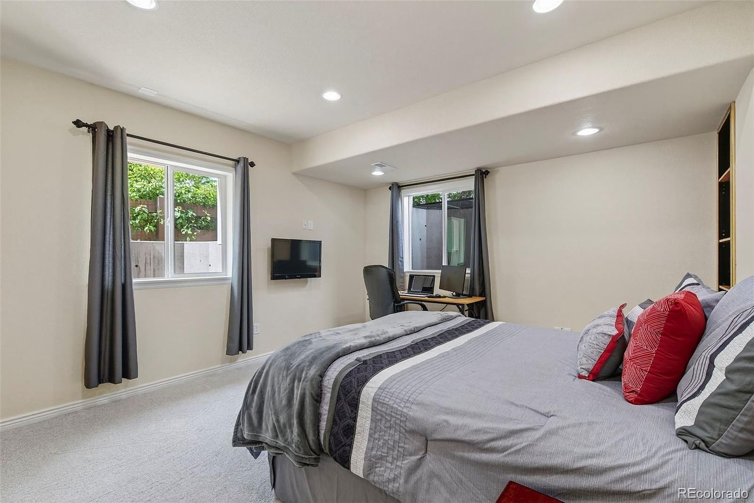 This is a cozy bedroom featuring a bed with gray bedding and red accent pillows. A desk with a computer setup sits near a window with gray curtains, and a wall-mounted TV is visible. The room has neutral-colored walls and carpet, creating a comfortable and functional space.
