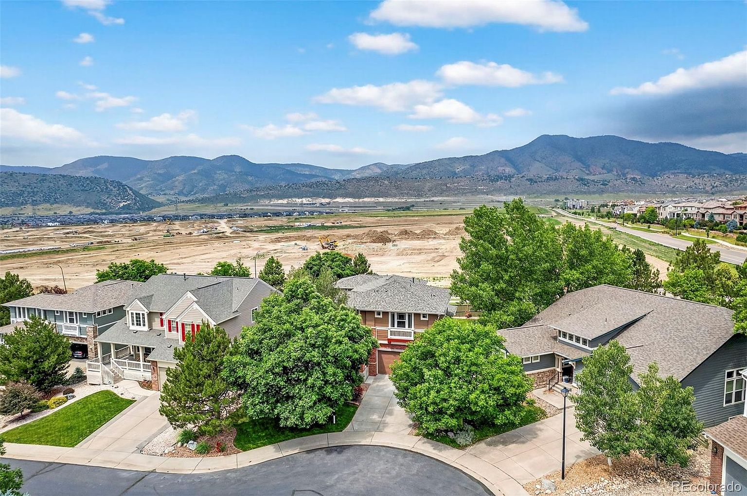 This aerial shot showcases a residential neighborhood with well-maintained homes, mature trees, and a cul-de-sac. In the background, there's a view of mountains and a large open area, possibly under development. The perspective gives a sense of the community's layout and its proximity to natural landscapes.