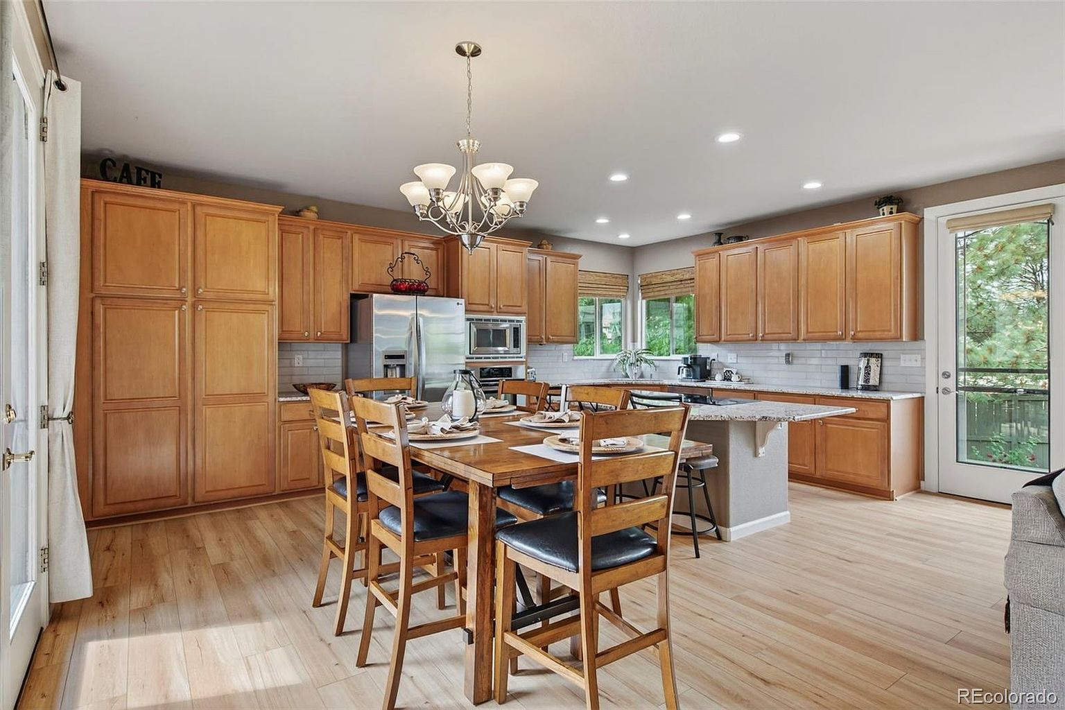 This is a well-lit kitchen and dining area featuring wooden cabinetry, stainless steel appliances, and a central island with a granite countertop. A wooden dining table with black leather chairs sits beneath a chandelier, creating a warm and inviting space. The hardwood floors and natural light enhance the room's appeal, making it perfect for family meals and entertaining.