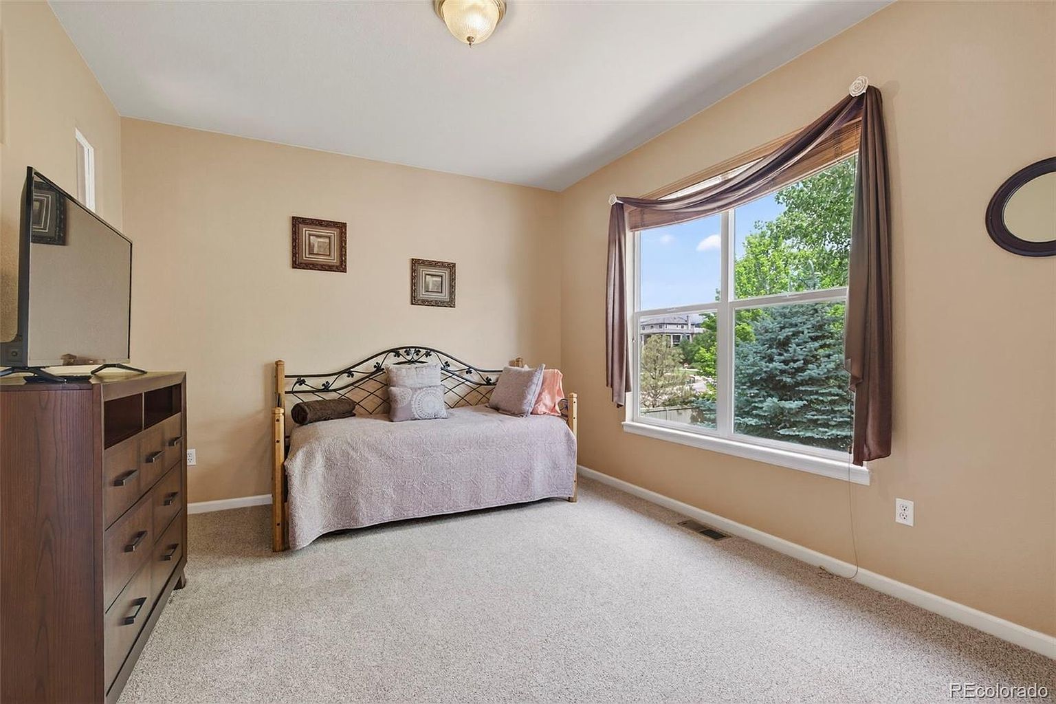 This is a cozy guest bedroom featuring a daybed with decorative pillows and a light-colored quilt. The room has neutral-toned walls, carpeted flooring, and a window with a view of greenery outside. A dark wood dresser with a television sits against one wall, and framed artwork adds a touch of personality.